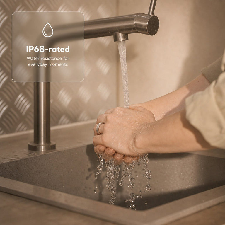 Hands wearing a silver smart ring under running water with an IP68-rated water-resistance note